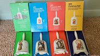 Four vintage Hummel Christmas collector bells along with their original boxes, arranged on carpet. Boxes are green, blue, red, and yellow and labelled with year and Christmas Bell name. Each bell features distinct figurine artwork in pastel colors with white handles.