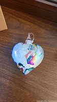 Heart-shaped glass ornament with multi-color iridescent surface, featuring abstract splashes of color and a hanging loop, photographed on a wooden surface.