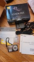 Nikon Coolpix S550 camera with original box, manuals, power cable, wrist strap, and battery laid out on a wooden surface.