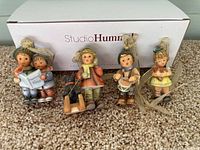 Group of five Studio Hummel porcelain Christmas tree ornaments displayed on carpet with white Studio Hummel box in background.