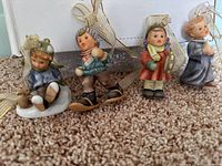 Four small Goebel Hummel ceramic figurine Christmas ornaments arranged on carpet, showing details and hanging ribbons.