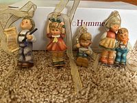 Photo of five Goebel Hummel Christmas figurine ornaments arranged on carpet with a Goebel Hummel box in background.