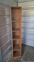 Full height frontal view of tall faux wood veneer bookcase shelf with five shelves visible, showing wood grain finish and slender profile.