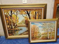 Photo showing two framed autumnal landscape paintings featuring fall foliage near water and trees