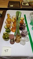 Full view of lot showing colored Pilgrim mini pitchers, tall slag glass swung vase, clear apothecary bottles, and wooden hourglass grouped on white surface.