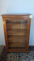 Front view of wooden cabinet with glass door revealing five shelves inside and carved decorative trim on top.