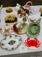 Photo showing assorted ceramic vessels, decorative plates, souvenir dishes, and figurines on a white tablecloth, including Maryland lobster dish and a Bavarian Turin plate.