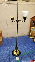 Full view of the brass and metal floor lamp standing against a white wall with a blue tarpaulin under it. Shows the white glass shade and brass bulb holder arms clearly.