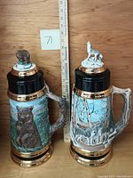 Two large ceramic steins with nature wildlife themes, one with bears and the other with wolves, both signed Linda Hogg, placed upright next to a measuring stick showing size.