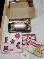 Vintage metal cookie press kit in original box with cylindrical press and nozzles, accompanied by pink vintage cookie cutters and original Easter-themed cookie cutter set box.