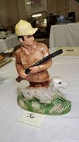 Full view of ceramic decanter showing hunter figure with dog on grassy base inside a room with other items.