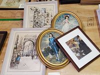 Five framed art prints arranged on a wooden surface including architectural sketches of Paris streets and classic portraits labeled as Blue Boy and Pink Girl.