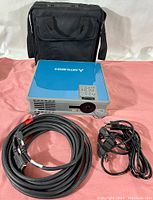 Projector placed in front of black carrying case with two cables laid in front: VGA and power cables, showing the front and side vents of the device.