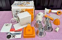 Photo showing the Moulinex Charlotte motor unit in white and orange with several metal and plastic attachments spread out on a purple surface, and the original box behind it.