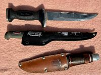 Three fixed blade knives displayed, two with sheaths; knives have black textured handles with metal guards, blades show wear and use.