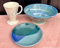 Photo showing all three items: tall beige mug, medium shallow bowl with blue-green glaze, large bowl with blue-green and pink design.