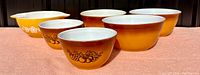 Overall view showing seven vintage Pyrex mixing bowls in yellow and amber-orange colors with distinct fruit and floral patterns.