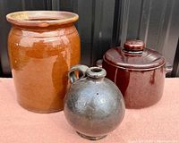 Photo showing all three stoneware items: one large brown crock, one smaller reddish-brown crock with lid, and one round dark glazed jug with handle.