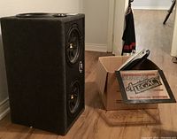 Photo of dual Kicker subwoofer box standing on floor next to a cardboard box containing peripheral items and an American Legacy sign.