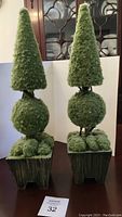 Pair of green topiary decorator pieces with three-tiered shapes, dark green planters on small wooden legs, shown on table in front of china cabinet