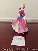 Side view of the Royal Doulton Windswept Melissa figurine in a pink and blue gown on a wooden surface with a MaxSold lot card.