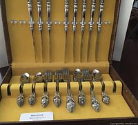 Full view of the complete Lyon stainless steel cutlery set arranged in yellow-lined brown box, including knives, forks, spoons, dessert forks, and teaspoons for 8 persons.