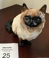 Front angle view showing the cat figurine's face, dark eyes, dark face, ears and tail, and cream body with fur texture detail.