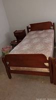 Photo of a single-size wooden bed frame with mattress in a room, showing the footboard and part of the frame structure.