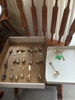 Complete set of 23 collectible souvenir spoons displayed in two cardboard trays on a wooden chair, showing different designs and metallic finishes.