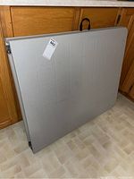 Folded 60x36 gray plastic folding table leaning against kitchen cabinets, with visible handle on top edge.