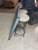 Photo showing side view of two metal frame bar stools with cream vinyl seats and folded card table propped upright showing metal frame and legs with pitting.