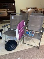 Photo showing four large folding lawn chairs with brown mesh seating, one smaller striped folding chair, black umbrella stand base on the floor, and three packages of webbing placed on chair seats.