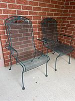 Pair of green metal lawn chairs with mesh backs and seats, decorative top backrest scrollwork, armrests, and four legs, positioned in front of a brick wall on a concrete porch.