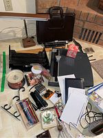 Photo showing a variety of office supplies arranged on a floor including a vintage desk lamp, black portfolio case, lined paper, alarm clock, pencils, plastic file trays, cutting board, and other small items.