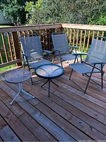 Three gray folding lawn chairs and two side tables on wooden deck. Chairs have metal frame and mesh fabric seats and backs. One small white metal slatted side table and one round glass top side table with metal legs shown.