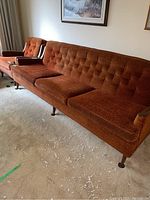 Photo showing the full burnt orange three-seater sofa with wooden legs and a matching armchair in the background.