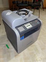 Front and side view of the Samsung SCX-5530FN multifunction printer showing the input tray, control panel, and power cord on top.