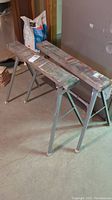 Two wood top saw horses with metal frames standing on the floor with paint and cuts visible on the tops.