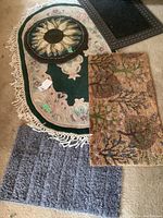Four mats on carpeted floor showing green floral carpet with fringe, round sunflower mat, multicolored floral door mat, and black rubber mat