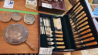 Overview of the Bakelite handle cutlery set in its case, showing multiple knives and forks, along with silver plate lined coasters and a silverplate butler with a wooden handle.