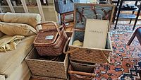 Photo showing overall view of several wicker and woven baskets including a picnic basket with open lid holding a book and cutlery.