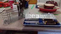 Carving set displayed with case, and metal wire stand for steak cutters on countertop with other kitchen items.