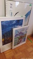 Three framed prints stacked leaning against a wall with varied sizes and different artwork styles.