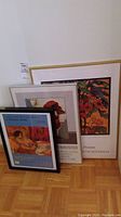 Three framed gallery advertising prints leaning against a wall on parquet floor. Prints include floral, abstract piano motif, and colorful painting.