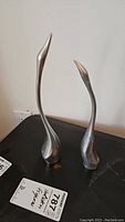 Pair of aluminum bird sculptures with elongated necks and smooth surfaces on a black surface, showing full figures.