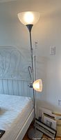 Photo of the larger floor lamp showing both the overhead light and the adjustable reading light against a white wall and near a white metal bed frame.