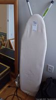 Ironing board with padded light cover and gray legs, leaning against a wall.