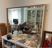 Large rectangular mirror with beveled edges and light wood frame, reflecting a room with furniture and decor. Mirror appears mounted to the wall with bolts.