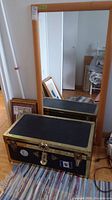 Framed mirror leaning against wall next to vintage black trunk with brass accents and lock, various materials inside the trunk.