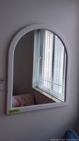 Arched white framed mirror hanging on wall reflecting a window and part of a bed
